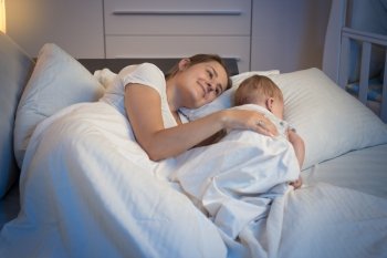 Night shot of happy smiling mother lying on bed with her beautiful baby