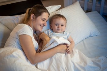Portrait of beautiful smiling woman lying in bed at night and hugging her baby boy