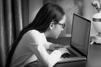 Closeup black and white portrait of teen girl with bad sight typing message on laptop