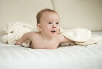 Funny portrait of surprised baby boy lying under blanket on bed