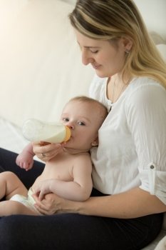 Toned portrait of young mother sitting on bed giving bottle to her baby