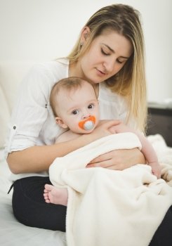 Toned portrait of young caring mother sitting on bed and holding baby on hands