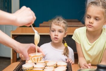 Two girls with a surprised look like mom pours batter into molds for cupcakes