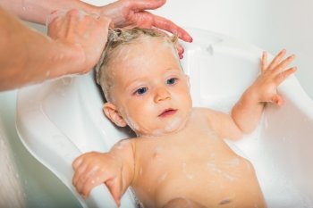 Little pretty wet baby boy in bath room with foam soap hair sitting and playing with mother's hand on white background, horizontal picture.