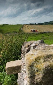 A hay baler at work in a field in the countryside, with a rock wall in the foreground