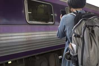 young woman holding mobile phone with backpack standing on platform at train station - travel concept