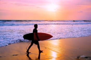 Surfer with surfboard walking on the sandy beach at sunset. Bali island, Indonesia