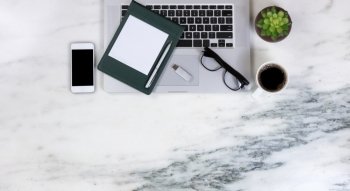 Flat lay of a partial laptop, plant, smartphone, pen, paper, reading glasses, thumb drive, and dark coffee on natural marble desktop