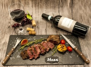Close-up of juicy beef steak striplon with a bottle and a glass of red wine on a black stone plate on a wooden table.. Close-up of juicy beef steak striplon with a bottle and a glass of red wine on a black stone plate on a wooden table