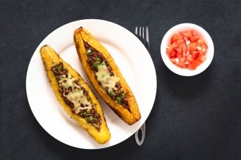 Baked ripe plantain stuffed with mincemeat, olive, green bell pepper and onion, sprinkled with cheese, a traditional dish in Central America called Canoa de Platano (Plantain Canoe), served with tomato and onion salad, photographed overhead on slate with natural light