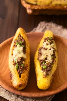 Baked ripe plantain stuffed with mincemeat, olive, green bell pepper and onion, sprinkled with cheese, a traditional dish in Central America called Canoa de Platano (Plantain Canoe), photographed on dark wood with natural light (Selective Focus, Focus one third into the image)