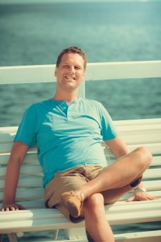 Handsome man tourist sitting on bench on pier. Guy enjoying summer travel vacation by sea. Fashion and relaxation.. Handsome man tourist on pier. Fashion summer.