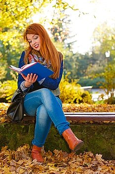 Nature, people, autumn concept. Ginger hair girl is reading book in park. Leaves are surrounding lady.