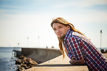 Man long hair relaxing by seaside . Man bearded long hair wearing plaid shirt casual style relaxing by seaside at summer sunny windy day 