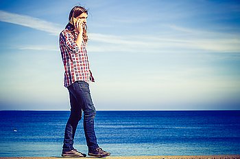 Man long haired casual style walking outdoor on stone wall receiving a call on his cell phone, guy using smartphone blue sky background