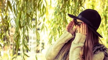 Adorable woman in sunglasses. Beauty and fashion of women. Young attractive fashionable girl wearing stylish hat waistcoat and sunglasses. Pretty woman around leaves of willow tree.