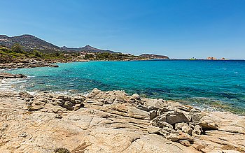 Aquamarine Mediterranean sea, rocks & maquis at Cala d’Olivu north of Ile Rousse in the Balagne region of Corsica with houses at Monticello in the background as well as a ferry in the distant harbour