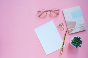 Flat lay of workspace desk, minimal stationery with note book, paper, pen, clip and eyeglasses on pink color background with copy space