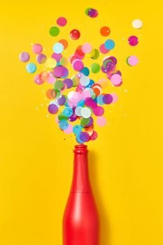 Holiday champagne red painted bottle with multicolored glitter as a bubbles foam on an yellow background, copy space. Flat lay.. Colorful confetti as a bubble foam from painted red wine bottle.