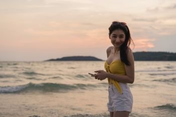 Asian teenage woman walking on beach. Beautiful girl using mobile phone checking social media near sea while enjoy her summer holiday vacation when sunset in evening.