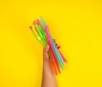 female hand holding multicolored plastic cocktail tubes, yellow background