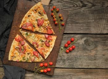 baked pizza with smoked sausages, mushrooms, tomatoes, cheese and dill, sliced food on a brown wooden board, close up