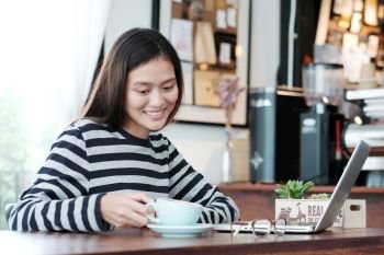 Young asian woman holding a coffee cup with smiling face, positive emotion while working with laptop computer at cafe background, lifestyle, people and technology