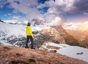 Young man in yellow jacket on the hill against snowy mountains at sunset in autumn. Landscape with sporty guy, rocks in snow, orange grass, blue sky with clouds. Travel in Italy in fall. Tourism