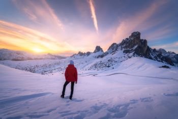 Young woman in snowy mountains at sunset in winter. Landscape with beautiful slim girl on the hill against snow covered rocks and colorful sky with clouds in the evening. Travel in Dolomites. Tourism.