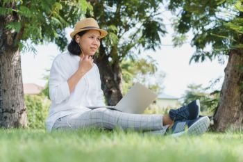 Asian women wear hat are using laptop with wired headphones while sitting on grass green  In the public park