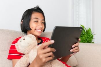 Asian little girl wearing a red shirt using tablet and smile While sitting on the sofa, Doll, at her home