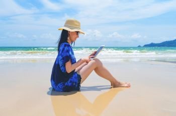 Happy Asian woman using a tablet at the beach during travel holidays vacation outdoors at ocean or nature sea at noon, Phuket, Thailand