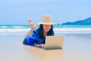 Happy Asian woman using a computer laptop at the beach during travel holidays vacation outdoors at ocean or nature sea at noon, Phuket, Thailand