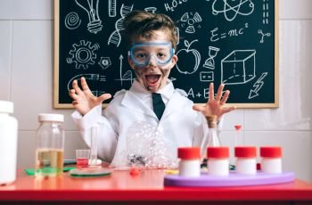 Portrait of happy little scientist with glasses and dirty face opening his arms behind of glass with soap foam over table. Happy little boy playing excited with experiment results