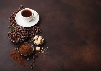 White cup of fresh raw organic coffee with beans and ground powder with cane sugar cubes with coffee tree leaf on dark background. Top view