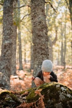 Little caucasian baby girl squatting wearing a wool cap in an autumn forest among ferns plays with plants