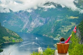 Female tourist enjoying scenic view over fjord Geirangerfjorden from Flydalsjuvet viewpoint, holding norwegian flag. Cruising vacation and travel.. Tourist over Geirangerfjord holds norwegian flag