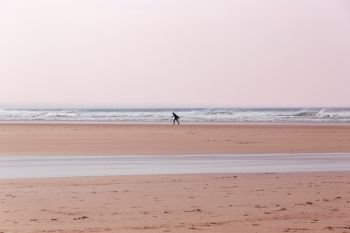 Lone surfer, carrying a surfboard, walking along the beach in Cornwall, UK