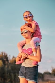 Sisters enjoying piggyback ride. Teenage girl carrying her younger sister up on the back and shoulders spending time playing together outdoors in the countryside. Candid people, real moments, authentic situations