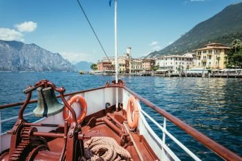 Bow of a boat with boat bell on a cruise tour. Blue water, mountain range and cute little village at Lago di Garda, Italy