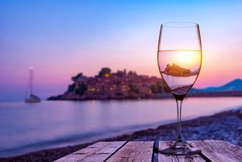 Wineglass on old wooden table on the beach of Sveti Stefan at sunset, Montenegro. Wineglass and Sveti Stefan