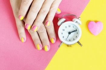 female hands with a bright pink-yellow gradient manicure on a yellow-pink background, a white alarm clock and a pink heart.

