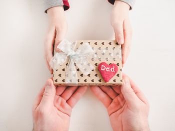 Child’s hands and adult man’s hands, beautiful gift box, ribbon and glazed cookies on a white, wooden background. Top view, close-up. Preparing for the holidays. Beautiful card for Women’s Equality Day. Close-up