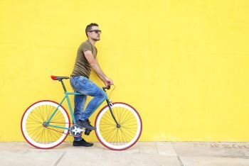 Side view of a young trendy man with a fixed bike wearing casual clothes while looking away against a yellow wall outdoors in a sunny day