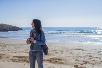 Front view of young woman wearing casual clothes standing on the beach while holding a camera and looking away in a bright day