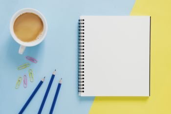 Office workplace minimal concept. Blank notebook, cup of coffee, pencil, paper clip on yellow and blue background. Top view with copy space, flat lay. Pastel color filter.