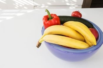Vegetables and fruit in a purple bowl on a white table closeup. Vegetables and fruit in a purple bowl on a white table