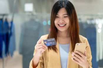 Portrait Asian woman using credit card with smart mobile phone for online shopping in department store over the clothes shop store background, technology money wallet and online payment concept