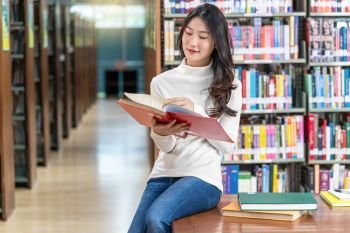 Asian young Student in casual suit reading the book in library of university or colleage on the wooden table over the book shelf background, Back to school concept