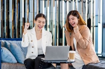 Two asian businesswomen hapiness celebrating together when checking success goal via technology laptop in modern office or coworking space, coffee break, relaxing and talking after working time